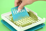 Hand using a powder spreader tool to spread green powder onto a capsule filling machine #color_teal
