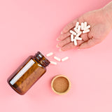 Hand holding white pills above an open brown glass bottle on a pink background