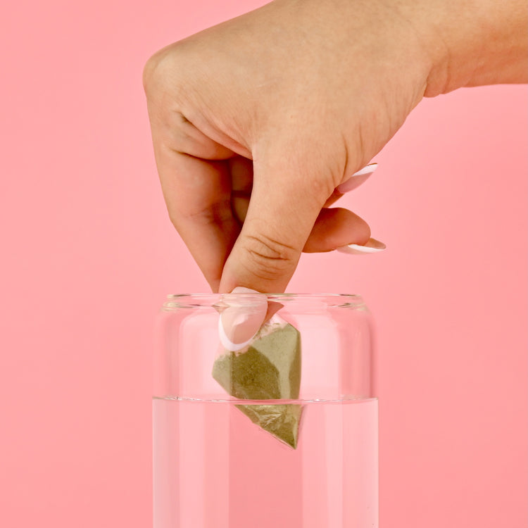 Hand placing a Blate Pape filled with green powder into a clear glass of water on a pink background